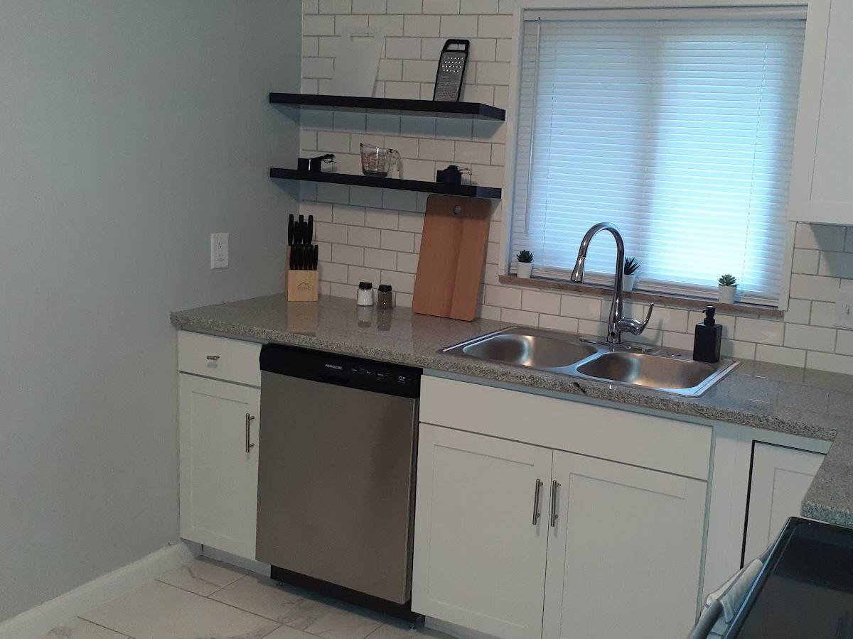 The same kitchen wall finished: gray speckled counters, a sink under the window with blinds, the black shelves stocked, and a stainless dishwasher.