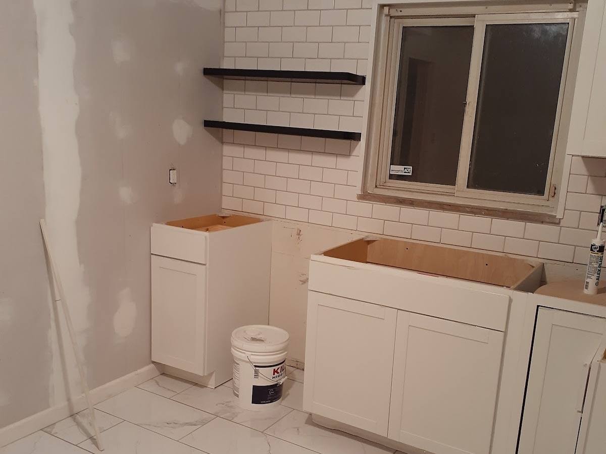 A kitchen wall with a sliding window, two black floating shelves and white subway tile, the white base cabinets set in but open on top with no counters yet.