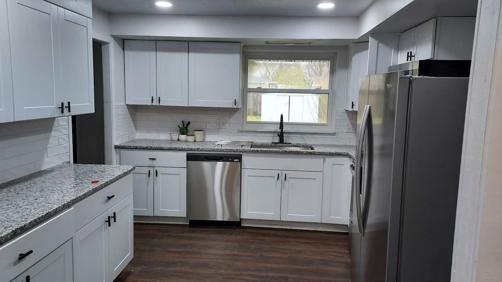 A finished kitchen with white shaker cabinets, speckled granite counters, a stainless dishwasher and refrigerator, and dark plank flooring.