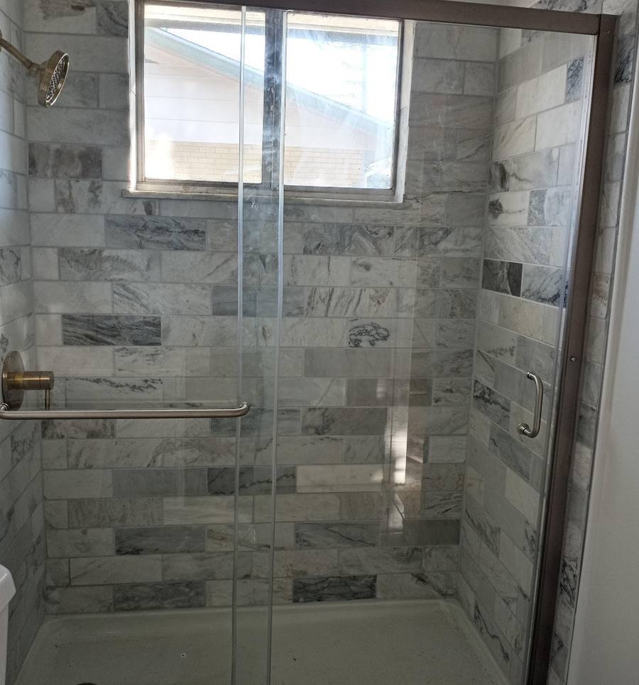A shower stall tiled in grey marble-veined rectangles with a brass grab bar, a small window and a sliding glass door.